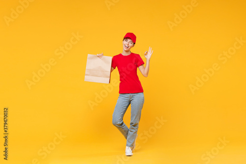 Full length body delivery employee woman in red cap blank t-shirt uniform work courier service in quarantine coronavirus covid-19 hold craft paper takeaway bag mockup isolated on yellow background.