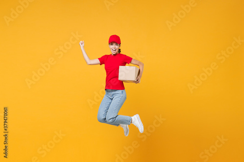 Full length body jumping delivery employee woman in red cap blank t-shirt uniform work courier in service during quarantine coronavirus covid-19 virus hold cardboard box isolated on yellow background.