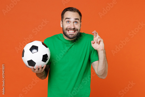 Man football fan in green t-shirt cheer up support favorite team with soccer ball wait for special moment keeping fingers crossed make wish isolated on orange background. People sport leisure concept. © ViDi Studio