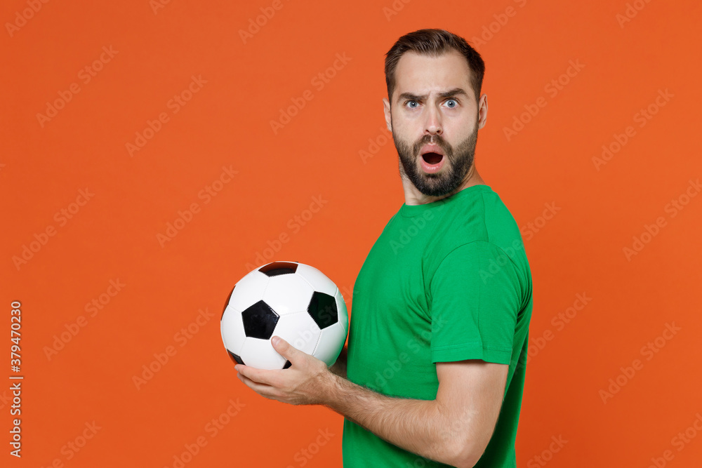 Side view worried young man football fan in green t-shirt cheer up support favorite team with soccer ball looking camera isolated on orange background studio. People sport leisure lifestyle concept. Side view worried young man football fan in green t-shirt cheer up support favorite team with soccer ball looking camera isolated on orange background studio. People sport leisure lifestyle concept.