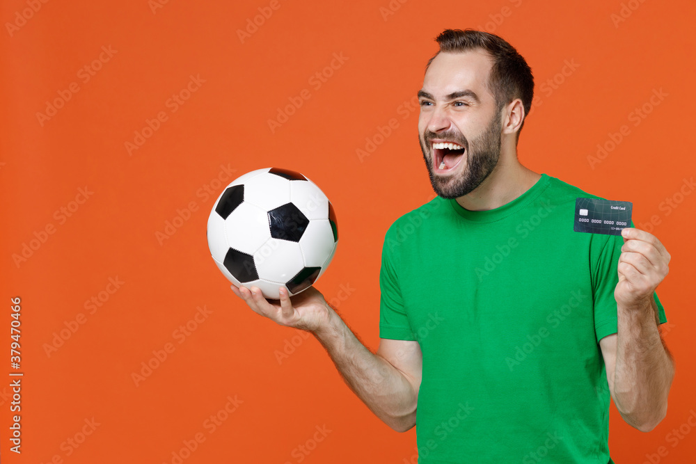 Cheerful young man football fan in green t-shirt cheer up support favorite team with soccer ball hold credit bank card isolated on orange background in studio. People sport leisure lifestyle concept. Cheerful young man football fan in green t-shirt cheer up support favorite team with soccer ball hold credit bank card isolated on orange background in studio. People sport leisure lifestyle concept.