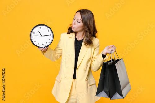 Shocked young brunette woman 20s wearing basic light suit jacket hold package bags with purchases after shopping clock looking aside isolated on yellow background, studio portrait. Black friday sale.