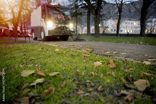 New generation of small electric street sweeper removing fallen leaves in body at autumn city park. Municipal urban services using ecology green vehicle lorry to clean streets from foliage. photo