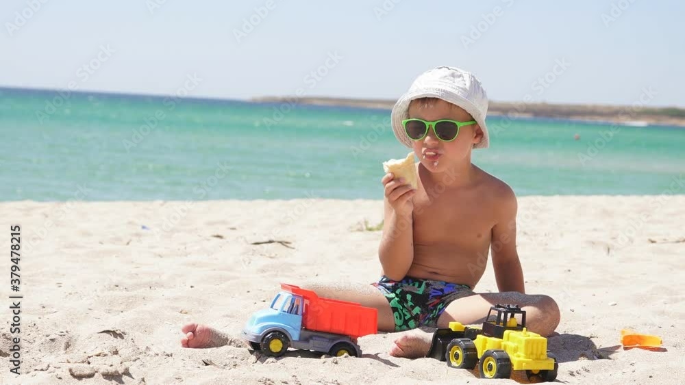 Tanned boy sits on a sandy beach and eats ice cream. Azure sea on a Sunny summer day on the horizon. Tanned boy sits on a sandy beach and eats ice cream. Azure sea on a Sunny summer day on the horizon.