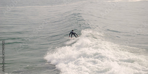 Surfista a surfar de pé em cima prancha na crista de uma onda do mar com um fato de neoprene vestido ao fim da tarde