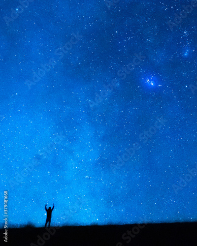 Person raising arms under the blue starry sky at night. Silhouette of a person observing the immensity of the universe, the stars, the constellations and the planets.