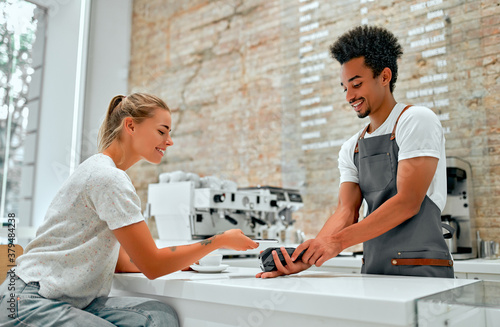 Female customer making payment through mobile phone at counter in cafe with young man. Barista holding credit card reading machine in front of female costumer with cell phone.
