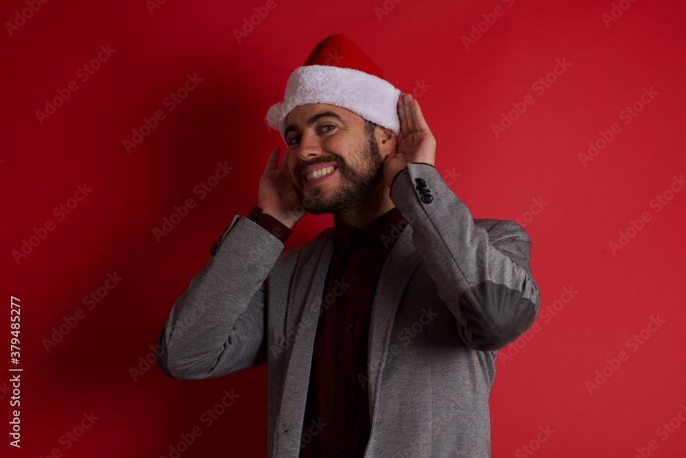 Young caucasian businessman wearing casual clothes and Santa hat standing over isolated red wall Trying to hear both hands on ear gesture, curious for gossip. Hearing problem, deaf Young caucasian businessman wearing casual clothes and Santa hat standing over isolated red wall Trying to hear both hands on ear gesture, curious for gossip. Hearing problem, deaf