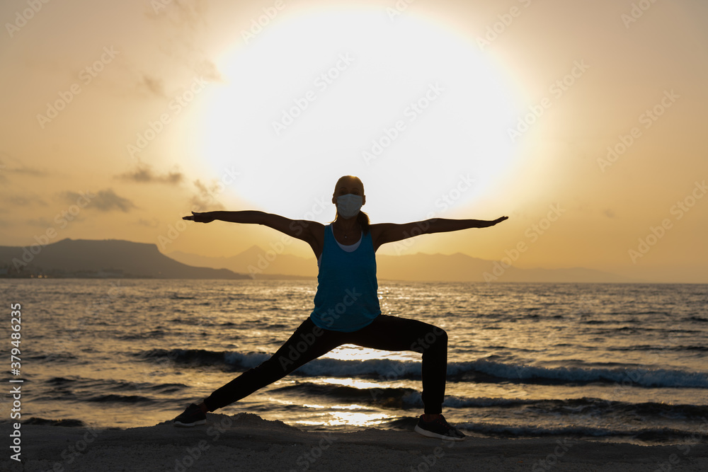 Silhouette, young woman with protective surgical face mask performs yoga stretching exercises at the beach at dusk during covid-19 coronavirus pandemic