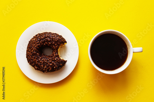 Breakfast concept. Morning coffee and bitten chocolate glazed donut with sprinkles on a bright yellow background. Baking, unhealthy food concept