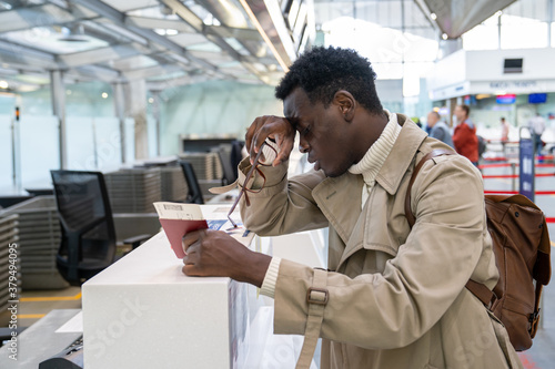 Black man upset about the cancellation flight. Afro-American traveler male overslept his flight, missed the plane, holding passport and boarding pass, standing at check-in counter at airport. 