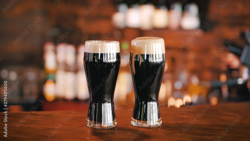 Two glasses of dark stout beer covered with foam, standing on the counter in a pub. Warm ambiente light of the bar