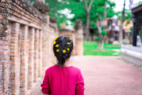 Back view of little asian girl in red dress with beautiful hair and yellow flower walking in the park