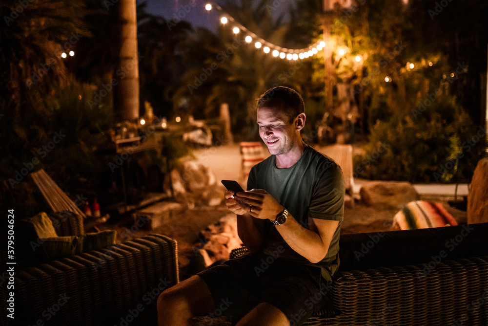 Smiling man using smart phone while sitting in garden during sunset Smiling man using smart phone while sitting in garden during sunset