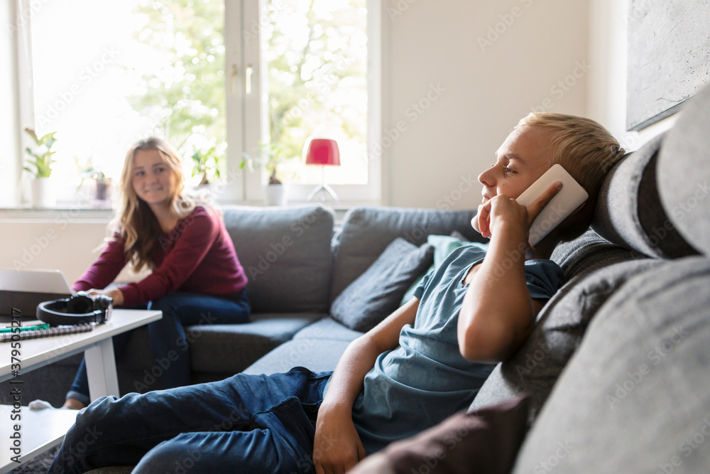 Male teenager talking on phone while smiling sister sitting on sofa at home Male teenager talking on phone while smiling sister sitting on sofa at home