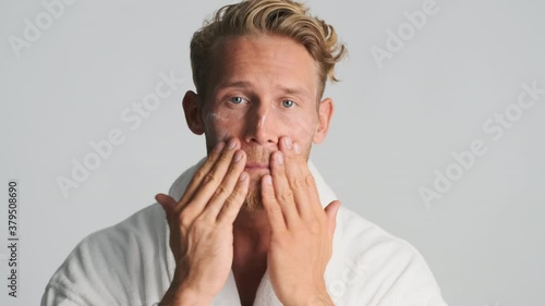 Attractive blond bearded man in bathrobe applying cream on face and smiling on camera over white background