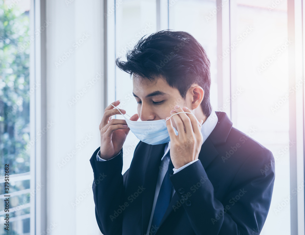 Young Asian businessman in suit wearing medical face mask in office. Young Asian businessman in suit wearing medical face mask in office.