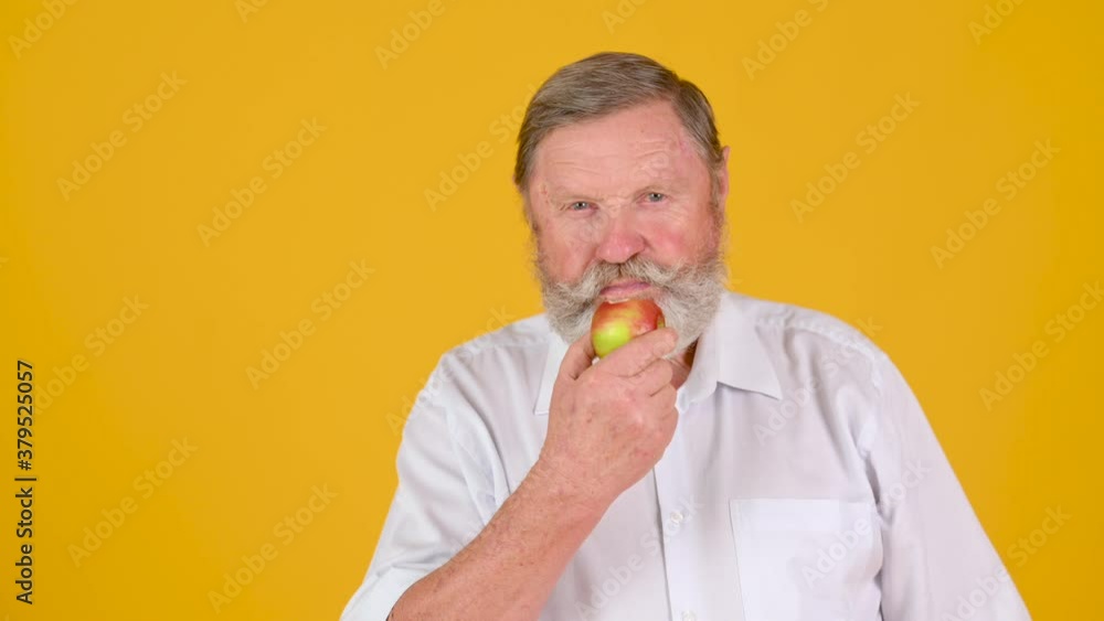 Elderly Gray-haired Man in White Shirt Eats Ripe Apple Isolated on Yellow Studio Background Elderly Gray-haired Man in White Shirt Eats Ripe Apple Isolated on Yellow Studio Background