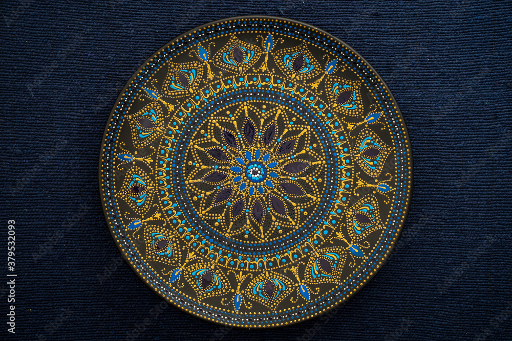 Decorative ceramic plate with black, blue and golden colors, painted plate on background of blue fabric, dot painting