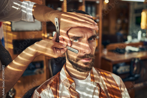 Serious young man having his beard groomed at a barbershop