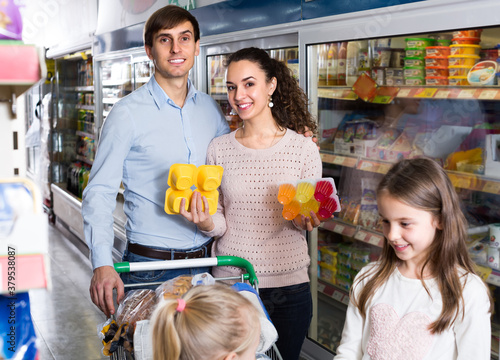 Happy young family with two little daughters purchasing yoghurts and desserts in supermarket. Focus on parents.