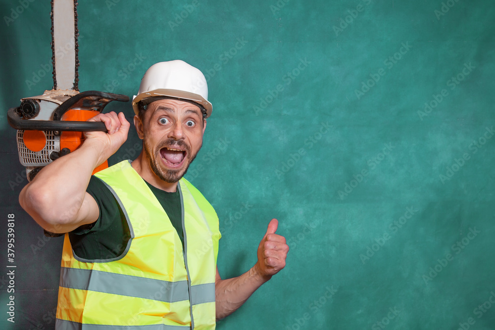 A joyful worker holds a chainsaw in his hand and shows a thumbs up gesture. A worker in a helmet and a signal vest. A bright emotion. A joyful worker holds a chainsaw in his hand and shows a thumbs up gesture. A worker in a helmet and a signal vest. A bright emotion.
