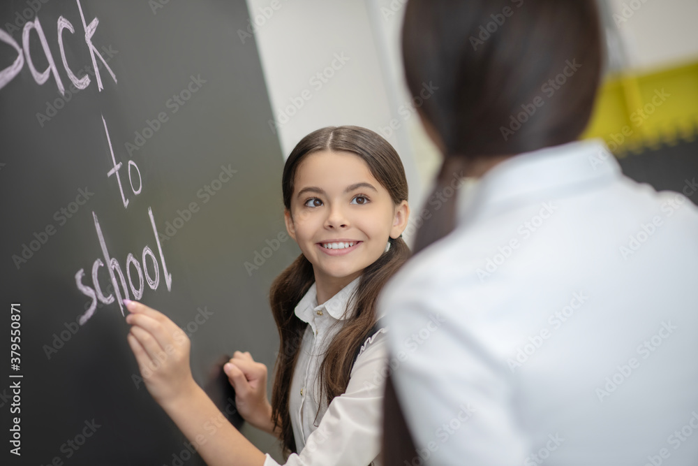 Smiling cute girl writing on the blackboard and looking at her teacher Smiling cute girl writing on the blackboard and looking at her teacher