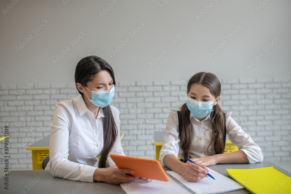 Teacher and schoolgirl in masks sitting in the classroom