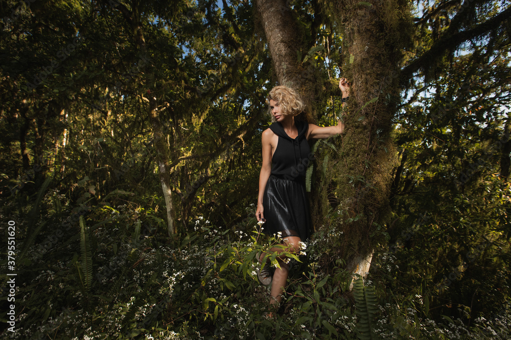  Beautiful blonde in a black dress with a hood stands near a tree in the forest. The girl got lost in the woods brooding dreamy Young women walking through the woods. Fashion style High quality photo