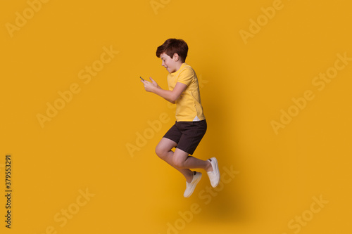 Ginger caucasian boy wearing a yellow t-shirt is jumping on studio wall while chatting on mobile