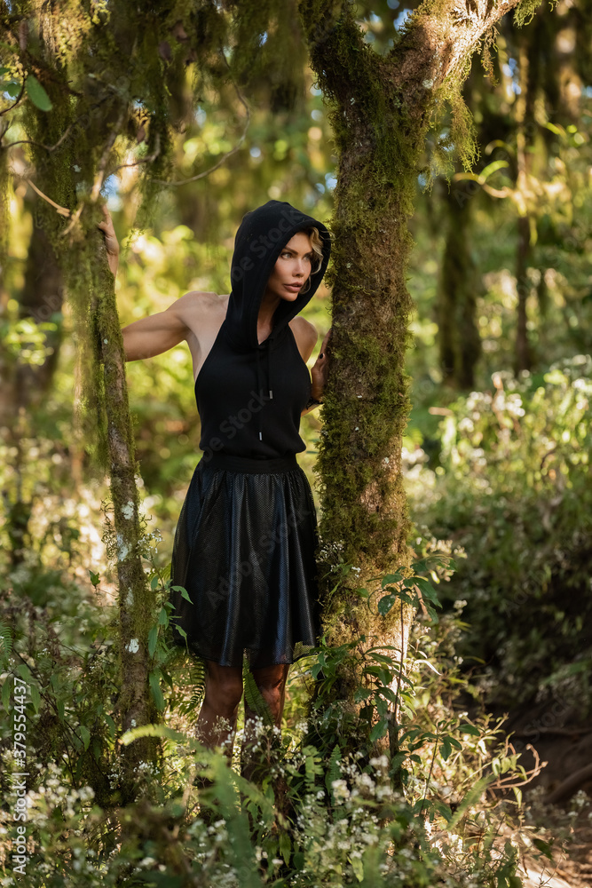 Brooding dreamy young girl blonde, black dress with a hood standing by a tree. The girl got lost in the woods. Trekking, fantasy, magical mystical forest. Girl standing by a tree. High quality photo Brooding dreamy young girl blonde, black dress with a hood standing by a tree. The girl got lost in the woods. Trekking, fantasy, magical mystical forest. Girl standing by a tree. High quality photo