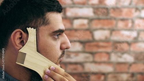 Bearded man showing template for shaving beard line in barber shop. Man using stencil for trimming bristle on face in barber salon