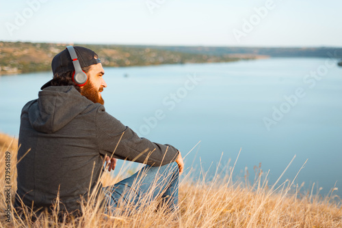 Positive bearded male sitting on the field listening to the music and looking away near a river © Vulp