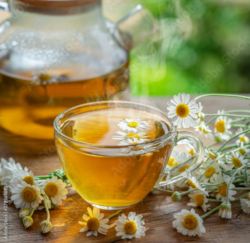 Herbal chamomile tea and chamomile flowers near teapot and tea glass on wooden table. Countryside background.