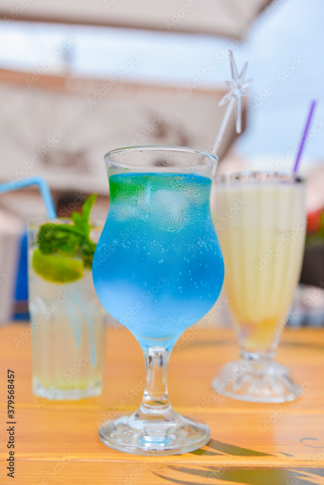 Three fresh juice drinks in tall glasses with plastic straws outside. Milkshake, blue curacao cocktail and mohito. Three fresh juice drinks in tall glasses with plastic straws outside. Milkshake, blue curacao cocktail and mohito.