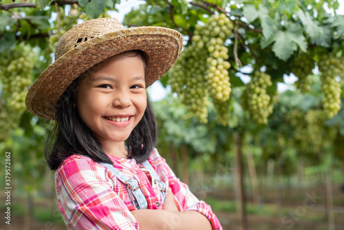 A cute girl in a hat and plaid shirt stood with her arms open in the garden, looking and smiling at the camera. The background is a vineyard. Asian children work hard to help the family business.