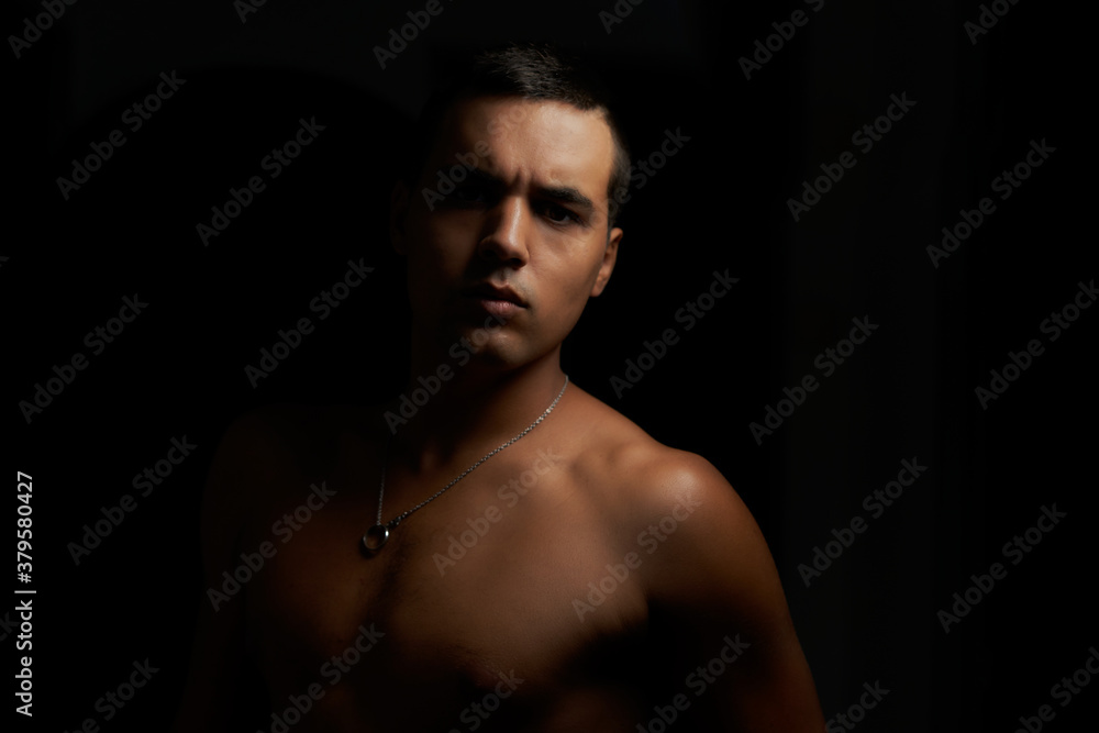 muscular young guy looking straight ahead. Shirtless muscular teenage boy on black background. Young shirtless brunette on black background. Athlete without a shirt and short hair with pendant.