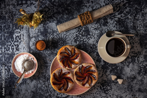 Small plum tarts on plate with cinnamon and icing sugar on the table with dark background, delicious puff pastry dessert with coffee black. photo