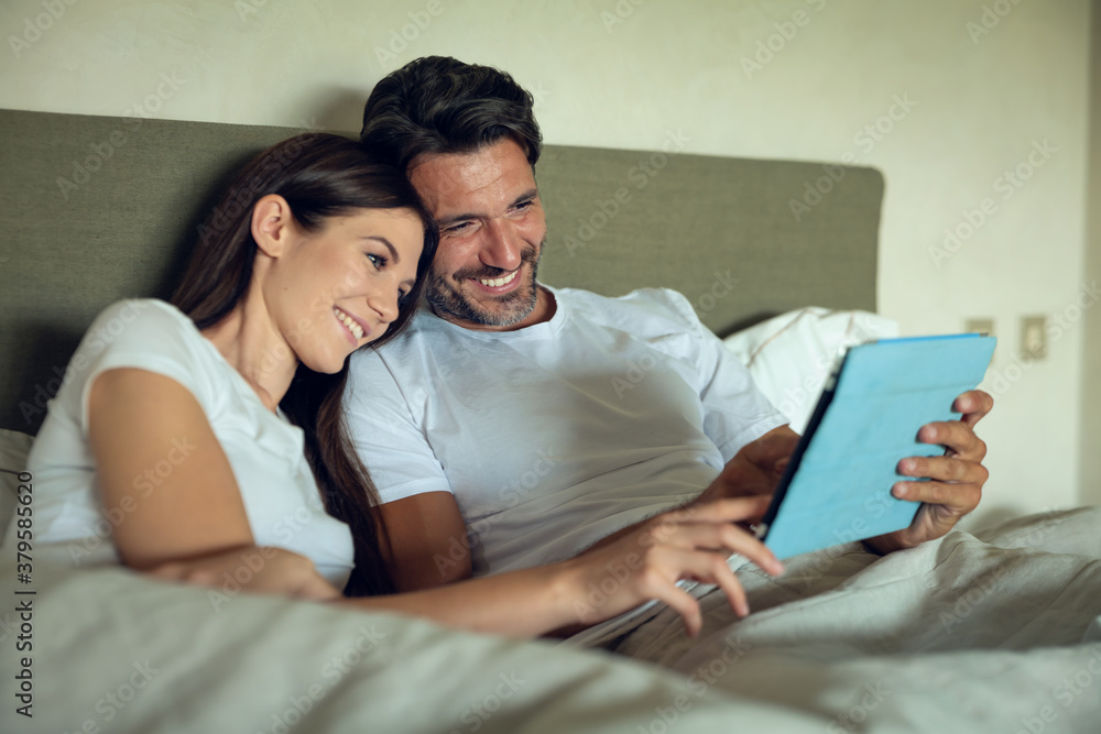 Authentic shot of young happy smiling just married couple in love is enjoying time together while using tablet for family entertainment on a bed in a bedroom just woke up in the morning in a sunny day