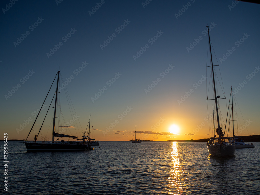Beach in the Formentera island with sailboats and yacht in the water anchored during sunset. Boats silhouette in the ocean.