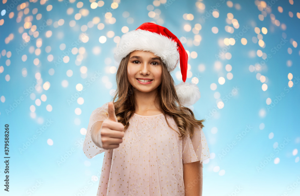 christmas, holidays and people concept - happy smiling teenage girl in santa helper hat showing thumbs up over festive lights on blue background christmas, holidays and people concept - happy smiling teenage girl in santa helper hat showing thumbs up over festive lights on blue background