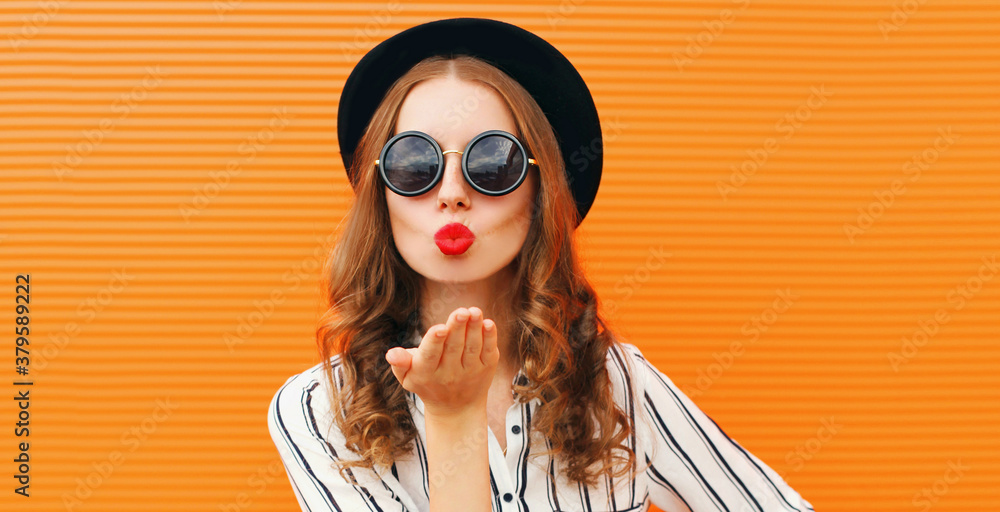 Portrait of stylish woman model blowing red lips sending sweet air kiss wearing a black round hat and striped white shirt on an orange background Portrait of stylish woman model blowing red lips sending sweet air kiss wearing a black round hat and striped white shirt on an orange background