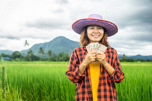 young Asian female famer hand holding Thai banknote money with happy face smiling over green rice farm 