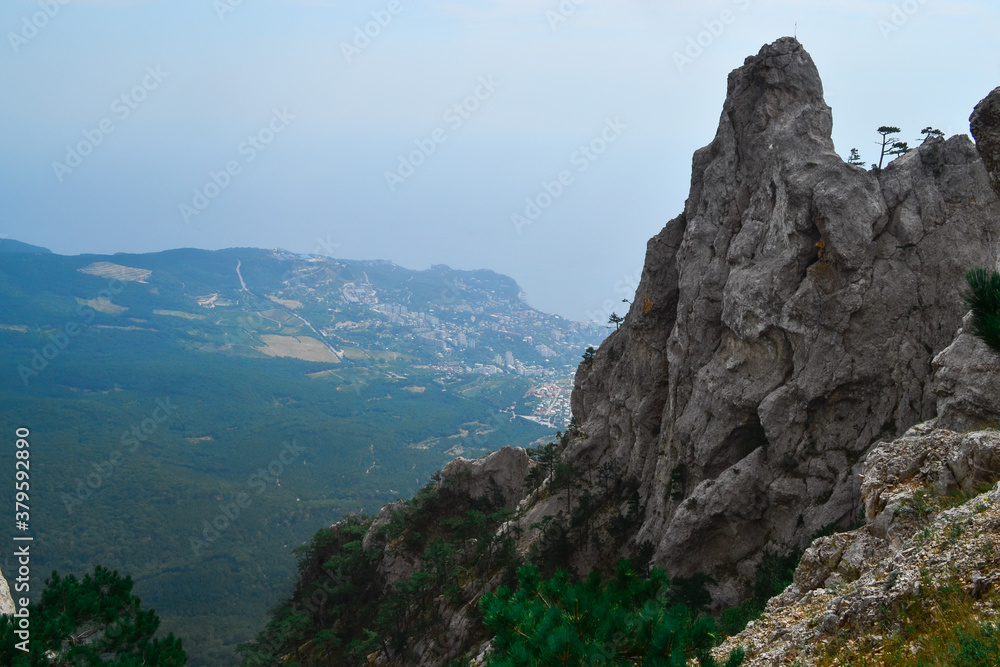 view of large stone rock among green trees stands on cliff against backdrop of Black sea coast in Crimea, city is below