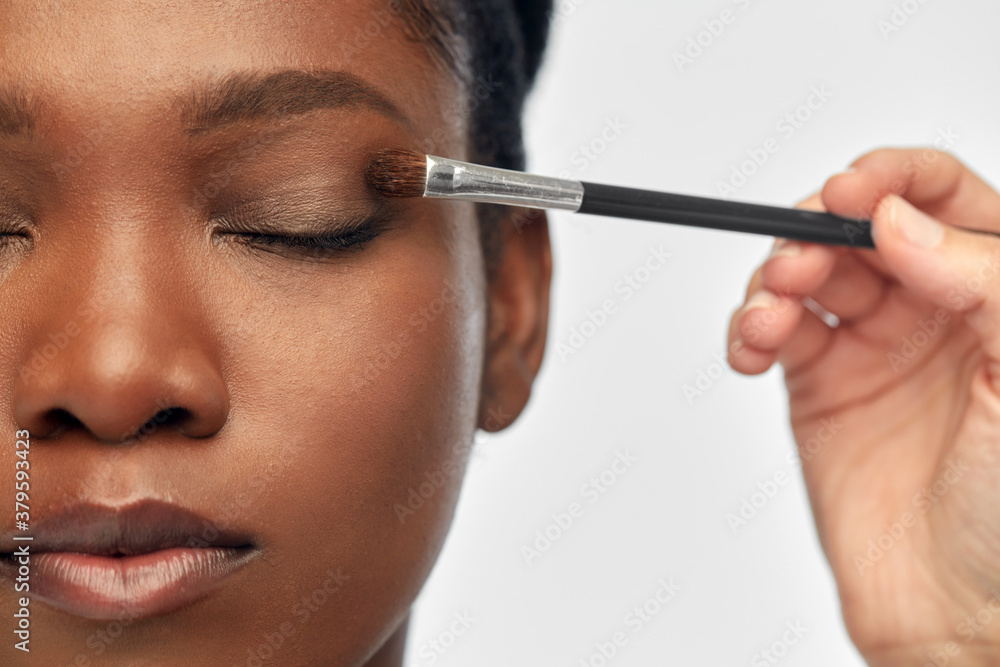 beauty, cosmetics and people concept - close up of face of beautiful young african american woman and hand with make up brush applying eye shadow over white background