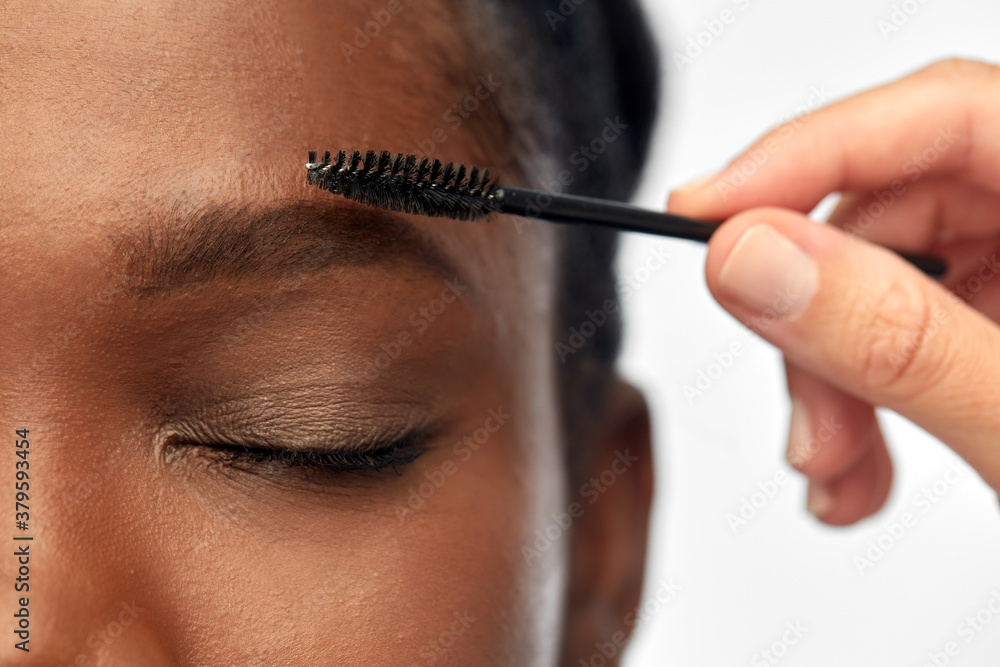 beauty, make up and cosmetics concept - close up of face of young african american woman and hand with mascara brush applying eyebrow shadows over white background