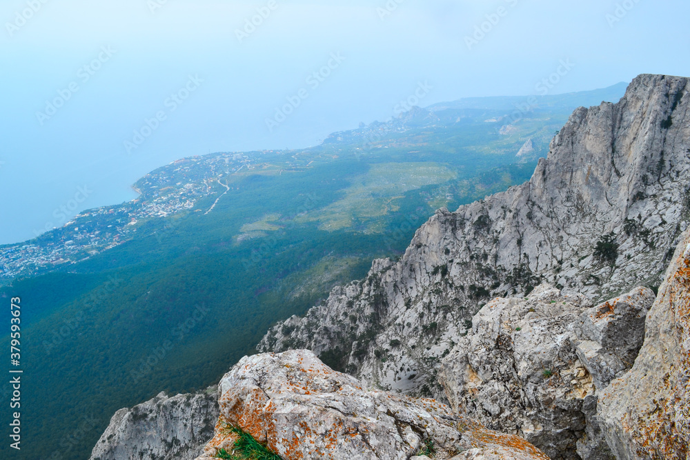 view of large steep stone rock cliff against the backdrop of blue sea coastline with city and forest