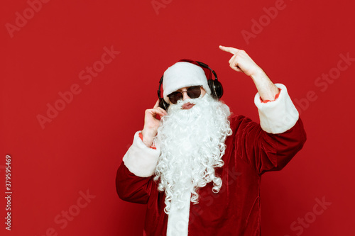 Studio portrait of cheerful Santa Claus in sunglasses listening to music in headphones and having fun on red background. Santa Claus partyman with headphones isolated on red background. X-mas