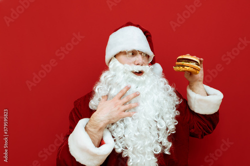 Portrait of hungry Santa Claus looking at burger in hand and stroking beard isolated red background.Funny man in Santa costume standing on red background with fast food and going to eat.Harmful food