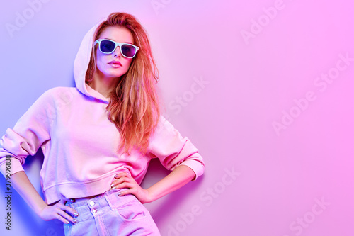 Fashionable hipster woman in Trendy outfit, stylish hair, makeup on pink purple neon light. Redhead model in hoodie, fashion jeans, sunglasses. Beautiful girl in autumn fall neon style © evgenij918
